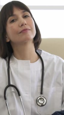 Kendine güvenen genç bayan doktor, klinikte sandalyeye oturmuş steteskopla kameraya bakarken yazıp gülümsüyor.