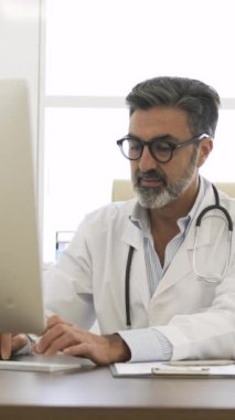 Steteskobu boynuna dolamış kendinden emin yetişkin bir erkek doktor. Klinikte otururken kameraya bakıyor ve konuşuyor.