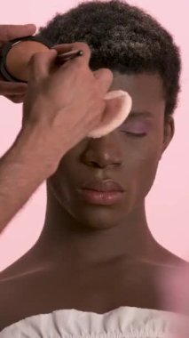 Hasat yoğunlaştırılmış makyaj sanatçısı pembe arka planda gözleri kapalı oturan Afro-Amerikan travestinin göz kapağına mor göz farı sürüyor. Dikey görüntüler