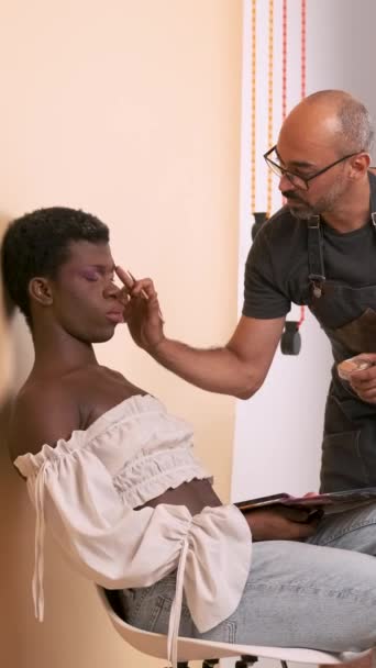 アフリカ系アメリカ人のドラッグクイーンの唇に口紅を塗る男性メイクアップアーティストの全長は ベージュの背景に対してスタジオの椅子に座っている間ピンクのトップを着用します バーティカル映像 — ストック動画