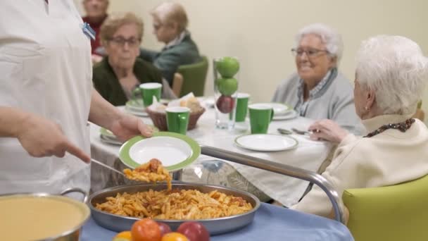 ラックは 退職した家の食堂でテーブルの周りに集まる高齢者のための美味しいマカロニにサービスを提供する匿名の女性労働者のゆっくりとした動きを集中させます — ストック動画