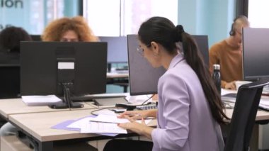 Bilgisayar masasındaki dokümanların verilerini analiz etmekle meşgul genç bir iş kadını ve çalışma alanındaki meslektaşları birlikte çalışıyor.