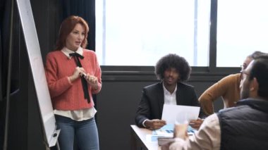 Kızıl saçlı kadın çalışanın, meslektaşlarına stratejilerini açıklarken ve konferans sırasında soruları cevaplarken beyaz tahtaya notlar aldığı gerçek zamanlı.