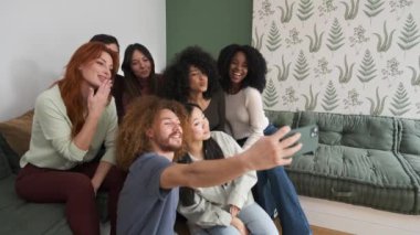 Neşeli, çoklu etnik çeşitliliğe sahip genç erkek ve kadın arkadaşlar, hafta sonu evde akıllı telefon kullanarak selfie çekiyorlar.