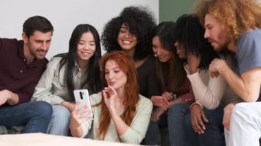 Gülümseyen kızıl saçlı kadının neşeli arkadaşlara cep telefonuyla fotoğraf göstermesi ve birlikte boş zaman geçirmesi.