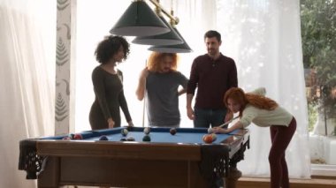 Çoklu etnik çeşitlilikteki erkek ve kadın arkadaşlar hafta sonu evde oyun odasında bilardo sopasıyla bilardo oynayan genç kadını izliyorlar.