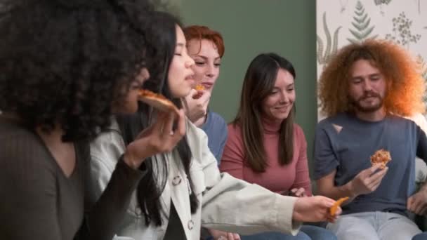 自宅で週末パーティー中にリビングルームで美味しいピザスライスとナチュオチップを食べながら話している多民族の若い男性と女性の友人のグループ — ストック動画