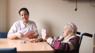 Beyaz üniformalı yetişkin bir dadının boş zamanlarında masada oturup tekerlekli sandalyeli yaşlı bir kadınla kart oynaması.