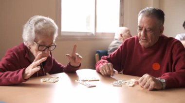 Yaşlı erkek ve kadın arkadaşlar günlük kıyafetleriyle masada domino oynarken huzurevinde vakit geçiriyorlar.