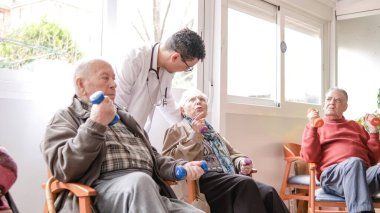 Dost canlısı erkek bakıcı rehabilitasyon merkezindeki fizyoterapi seansında yaşlı insanlara dambılları kaldırmada yardımcı oluyor.