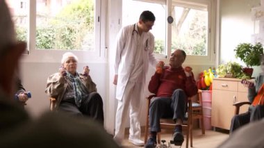 Genç bir doktorun, rehabilitasyon merkezinde bisiklet pedalları kullanarak ve dambılları kaldırarak yaşlı bir adamla konuşması.