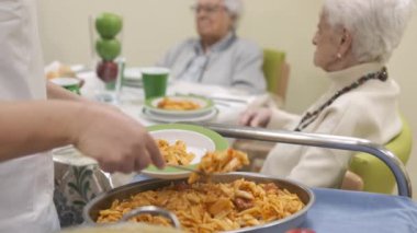 Huzurevinin kantininde toplanan yaşlılar için lezzetli makarna servisi yapan isimsiz kadın işçinin yavaş çekimde odaklanmasını sağlayın.
