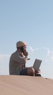 Erkek gezgin kum tepesinde dizüstü bilgisayar kullanarak oturuyor. Dijital göçebe ya da uzaktan çalışan kavramı