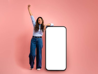 Model kadın mutlu bir şekilde poz veriyor, kopyalamak için boş ekranı olan dev bir cep telefonuna yaslanıyor, pembe arka planda..