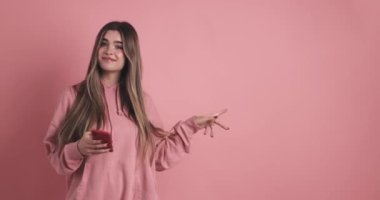 Uzun saçlı, sıradan kıyafetlerle gülümseyen, akıllı telefon kullanan ve pembe arka planda duran genç bir kadın.