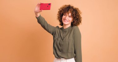 Pembe elbiseli, kıvırcık dudaklı, gülümseyen genç bir kadın. Bej arka planda dururken akıllı telefonuyla selfie çekiyor.