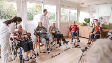 Üniformalı mutlu bakıcılar rehabilitasyon merkezindeki fizik tedavi seansında pedal egzersizi yapanlara yardım ediyor ve dambılları kaldırıyor.