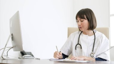 Steteskopla boynuna reçete yazıp hastanede bilgisayarla masada oturan kendinden emin genç bir kadın doktor.