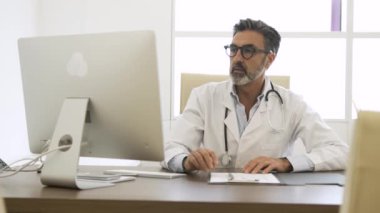 Masaüstü bilgisayarı kullanarak steteskopu boynuna dolamış ve klinikteki sandalyede otururken kameraya bakan mutlu olgun bir erkek doktor.