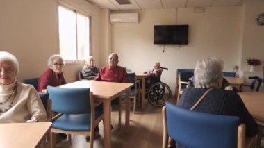 Huzurevi kantinindeki masalarda oturan yaşlı erkek ve kadınlar rehabilitasyon merkezinde kameraya bakıyor.