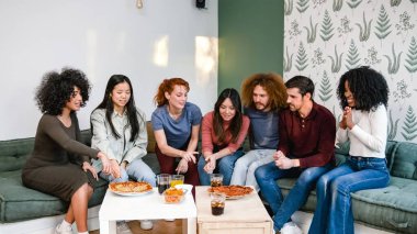 Bir grup etnik çeşitliliğe sahip erkek ve kadın arkadaş hafta sonu partisinde oturma odasında lezzetli pizzalar ve içecekler hakkında tartışıyorlar.