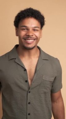 Heyecanlı Brezilyalı genç erkek afro saçlarını sıkarak kutlama yaparken bej arka planda duran kameraya bakıyor.