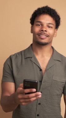 Tişörtlü, afro saçlı, cep telefonlu ve bej arka planda dikilirken kameraya bakan Brezilyalı bir erkeğin portresi.