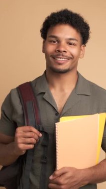 Sırt çantası ve bej arkaplan üzerine kitap tutarken kameraya bakan afro saçlı Brezilyalı genç üniversite öğrencisi.