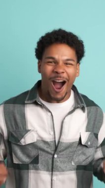 Heyecanlı Brezilyalı genç erkek, afro saçlı, yumruklarını sıkmış, başarısını kutlarken mavi arka planda duran kameraya bakıyor.