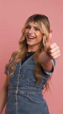 Kot elbiseli hoşnut bir transseksüel kadın kameraya bakıyor ve pembe arka planda dikilirken başparmağını kaldırıyor.