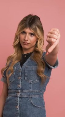 Kot elbiseli hoşnutsuz bir transseksüel kadın kameraya bakıyor ve pembe arka planda dikilirken başparmağını kaldırıyor.