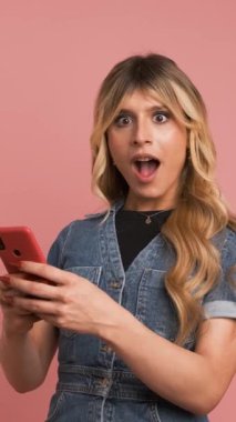 Kot pantolon giymiş, akıllı telefon kullanan ve pembe arka planda güzel haberleri kutlayan heyecanlı transseksüel kadın.