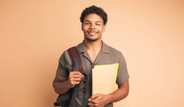 Sırt çantası ve bej arkaplan üzerine kitap tutarken kameraya bakan afro saçlı Brezilyalı genç üniversite öğrencisi.