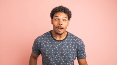 Tişörtlü afro-amerikalı adam pembe arka planda beklenmedik bir olayın olumlu sonucunu kutluyor.