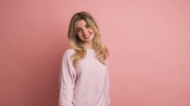 Pembe kazaklı, uzun sarı saçlı, kameraya gülümseyen ve pembe arka planda başını sallayan genç kadın modelin portresi.