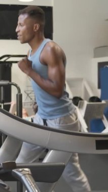 Sportif, genç, formda, spor giyimli, spor kıyafetli, modern spor salonunda antrenman yaparken koşu bandında egzersiz yapan Afro-Amerikan erkek sporcu.