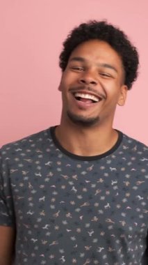 Tişörtlü genç Afro-Amerikalı adam pembe arka planda kameraya bakarken komik şakalara gülüyor.