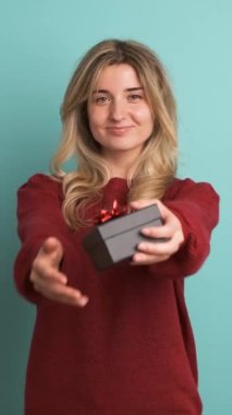 Sarı saçlı, kahverengi kazaklı, gülümseyen genç bir kadının portresi hediye kutusu veriyor ve mavi arka planda dikilirken kameraya bakıyor.