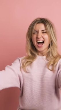 Stüdyo videosu ağır çekimde pembe arka planda genç bir sarışının enerjik bir şekilde yumruklarını kaldırmasını kutluyor.