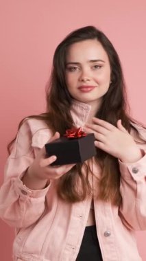 Pembe kot ceketli, uzun kahverengi saçlı, gülümseyen genç kadın portresi pembe arka planda sürpriz hediye paketi alıyor.