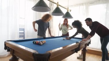 Modern ışık odasındaki farklı arkadaşlarla bilardo oynarken Afro saçlı genç bir kadının bilardo sopasıyla topa vuruşunun yavaş çekimi.