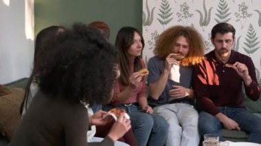 Hafta sonu evde parti sırasında oturma odasında sohbet ederken lezzetli pizza dilimleri yiyen çok ırklı erkek ve kadın arkadaşlar grubu.