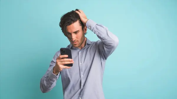 stock image Sad Caucasian male in shirt with black hair shaking head while using smartphone feeling upset about losing lottery while standing against blue background