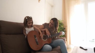 Evdeki modern oturma odasında boş vakitlerini geçirirken gündelik kıyafetleriyle kanepede gitar çalan Latin anne ve tatlı kızın yavaş çekimleri.
