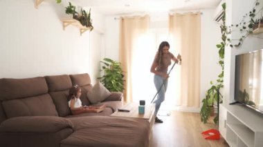 Uzun süpürge kullanarak yerleri süpüren ve modern dairenin oturma odasındaki akıllı telefonu kullanan küçük kızı izleyen mutlu Latin anne yavaş hareket ediyor.