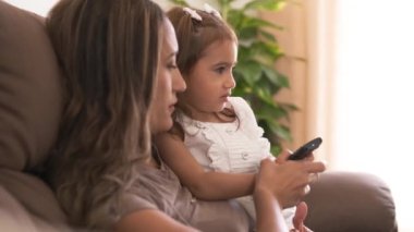 Oturma odasındaki kanepede televizyon izlerken uzaktan kumandayla kanal değiştiren Latin anne ve kızın yavaş çekim görüntüsü.