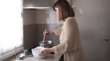 Mutfakta bir kadının masada servis yapmak için tencereden beyaz bir seramik kaseye et ve sos transferi videosu.