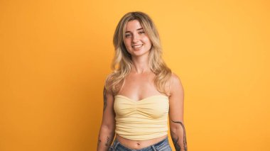 Gerçek zamanlı gülümseyen genç sarışın kadın kameraya bakarken sarı duvarın yanında durup başını onaylayarak ve vücudunu yanlara doğru hareket ettirerek eğleniyor.