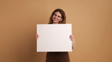 Gerçek zamanlı gülümseyen sarışın kadın kameraya eğik kafayla bakarken ayakta durup işaret parmağıyla kahverengi arka plandaki beyaz boş sayfayı işaret ediyor.