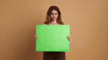 Yeşil paneli tutan endişeli ve üzgün genç bir kadının bej renkli arka planına sahip stüdyo videosu.